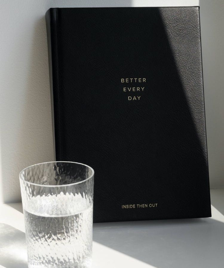 The Better Every Day Journal is placed upright against a wall with a glass of water placed in front of it